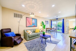 Un salon comprenant un canapé, un fauteuil et une table basse, un coin repas avec une table et des chaises, et une grande fenêtre avec des rideaux.