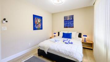 A bedroom with a bed, two bedside tables, a nightstand, a wall-mounted light fixture, and two paintings.