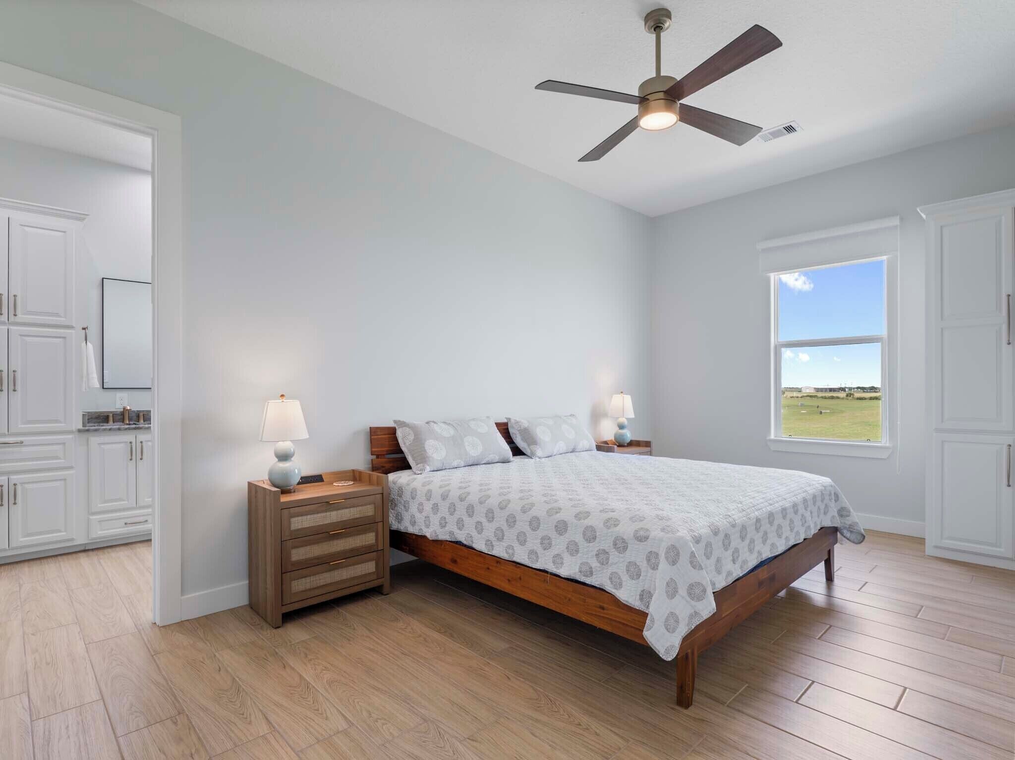 A bedroom with a wooden bed, two bedside tables with lamps, a ceiling fan, and a window with a view of a field.