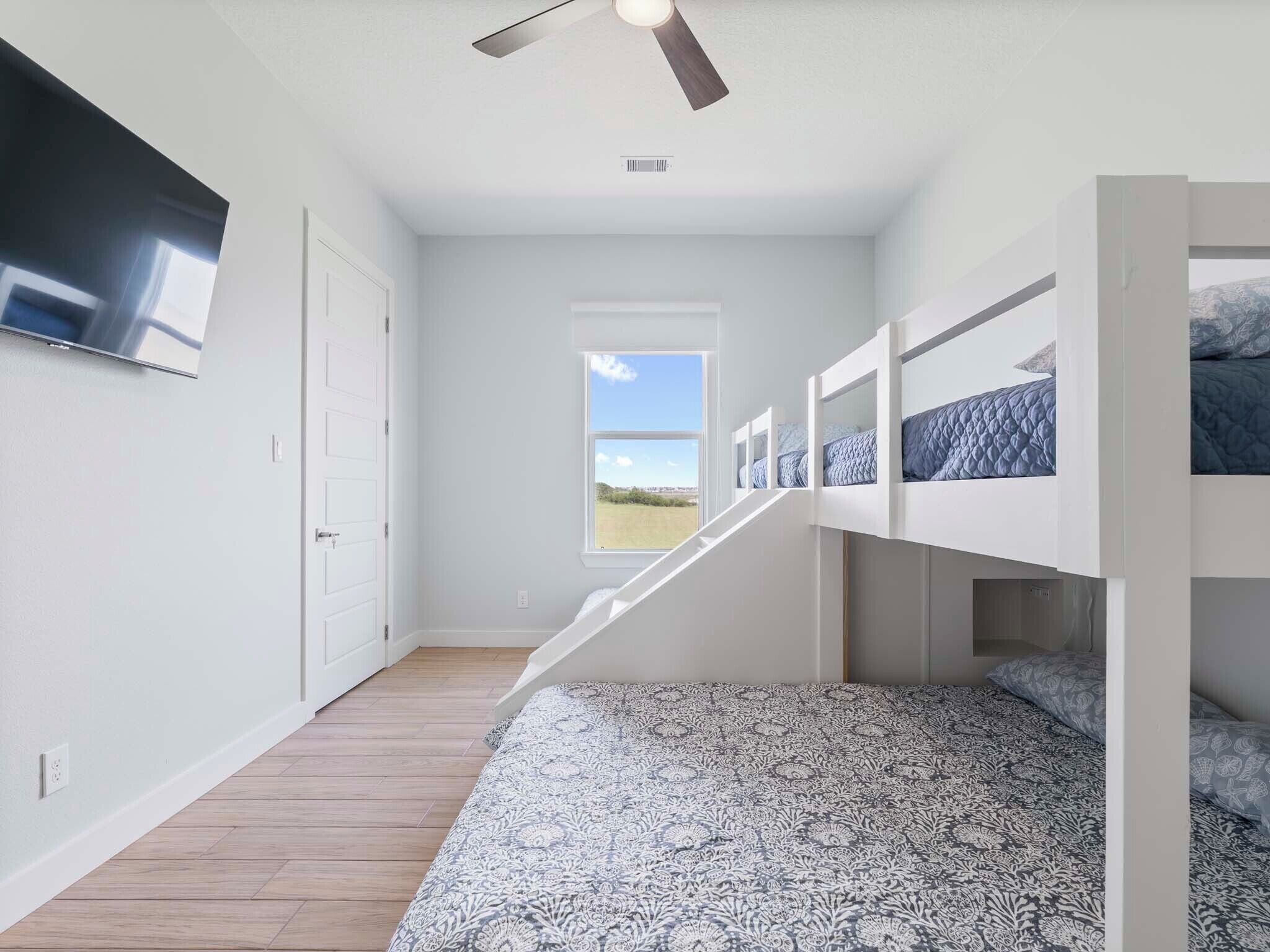 A bedroom with bunk beds, a ceiling fan, a TV mounted on the wall, and a window with a view of the outdoors.