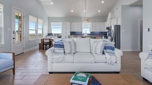 A spacious living room with a white sofa, wooden flooring, and a view of the kitchen. - Unwind on the Bay- Fishing, Pool, Tennis, Basketball courts & Kids' Playground (Palacios)