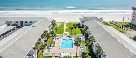 A hotel with a swimming pool surrounded by palm trees, a beach view, and a parking area.