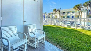 Un espace extérieur abrité, aménagé avec des chaises blanches et un banc, une clôture blanche et une pelouse bien entretenue agrémentée de palmiers.