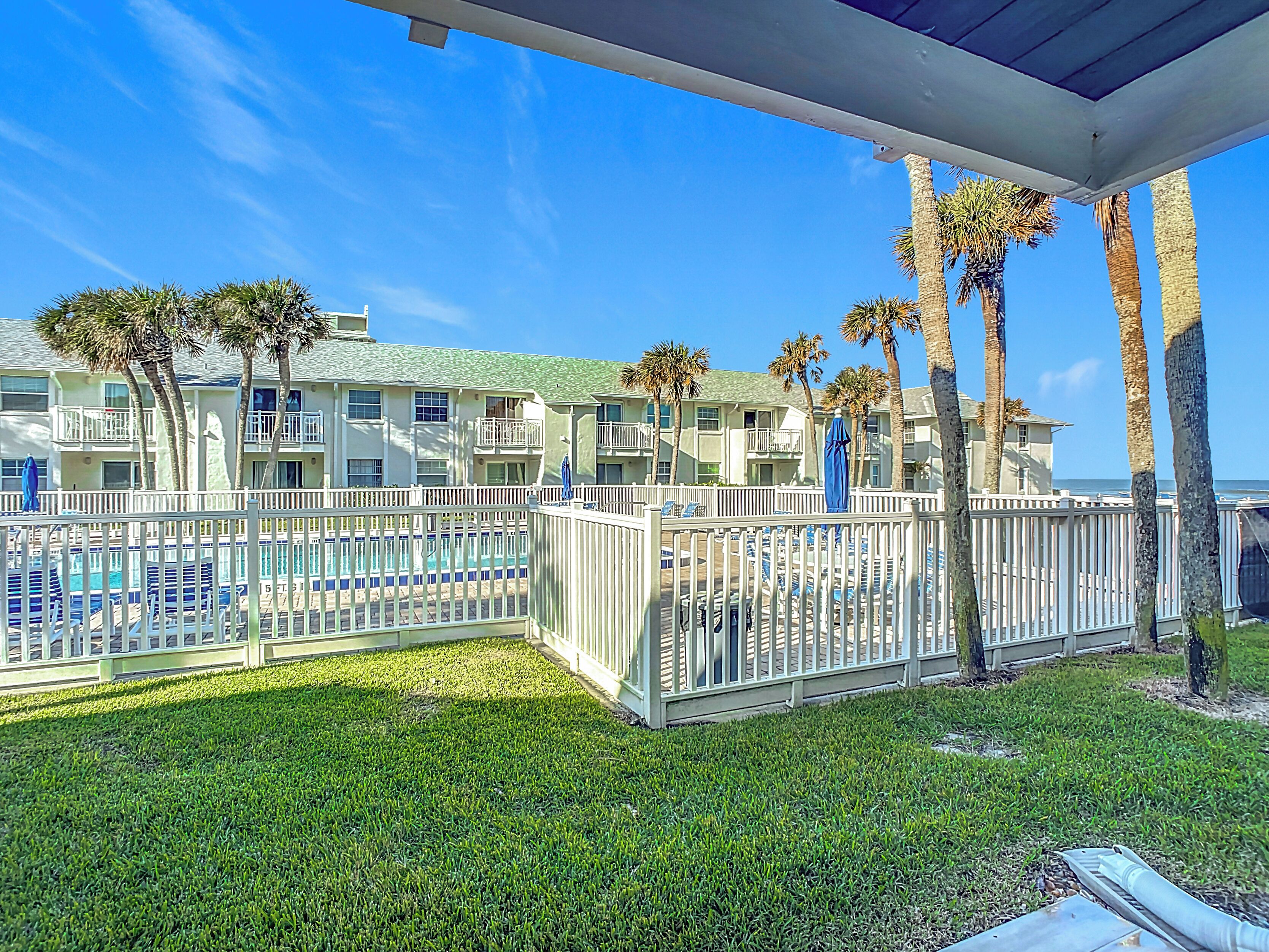 A pool area with a white fence, palm trees, and a building with balconies in the background.