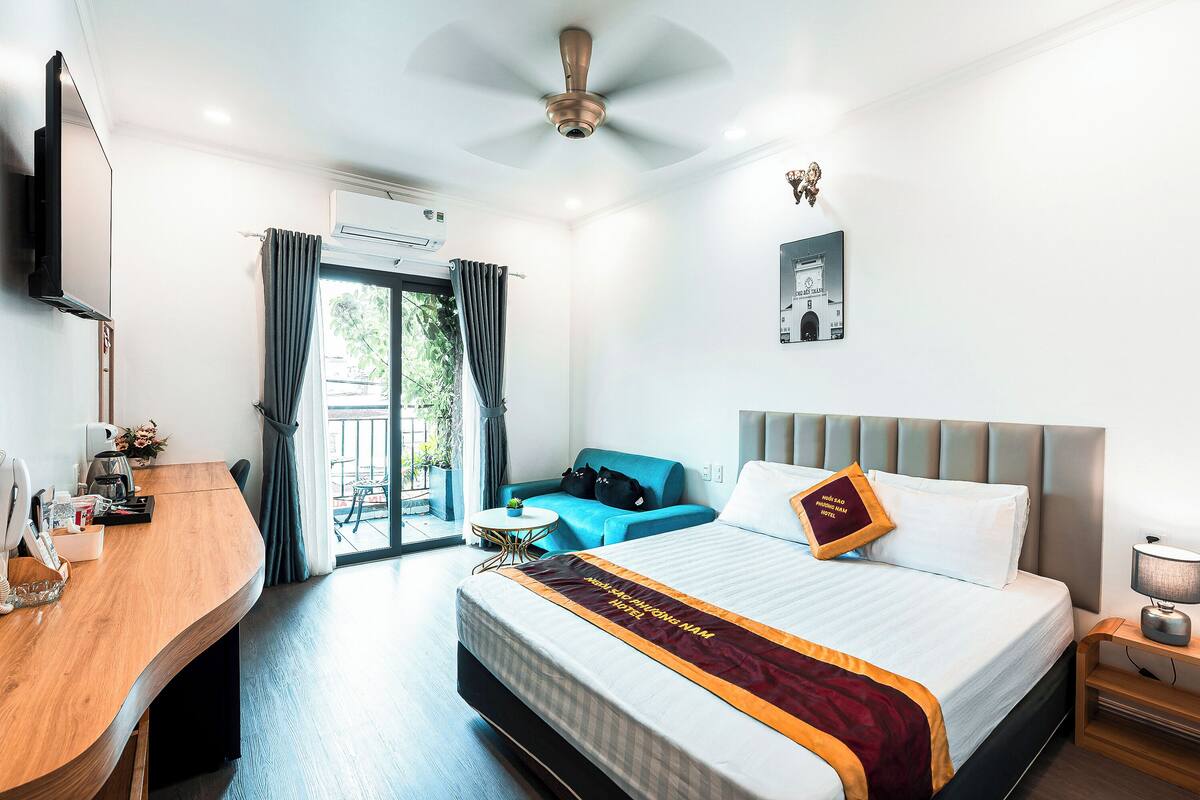 Habitación de hotel con cama, escritorio, silla, una mesita, ventilador de techo y ventana con cortinas.