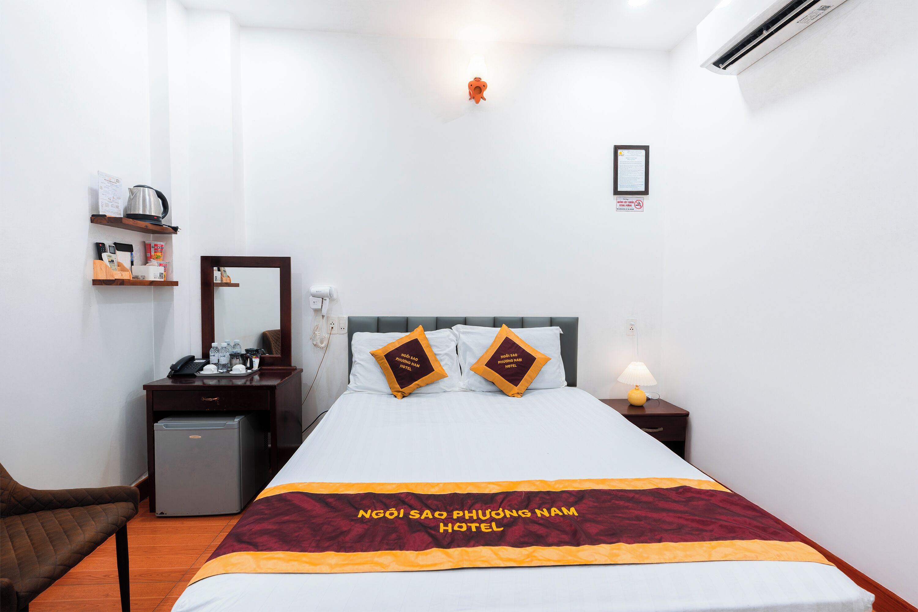 Habitación de hotel con cama, escritorio, silla, refrigerador pequeño, mesita de noche con lámpara y aire acondicionado de pared.