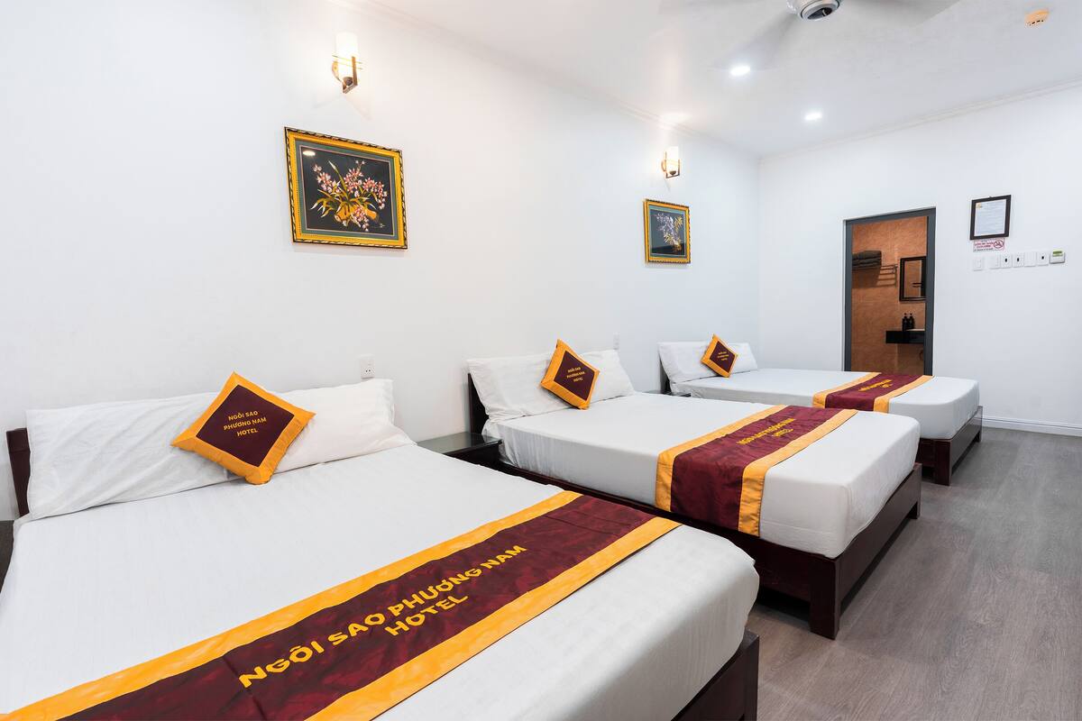 Habitación de hotel con tres camas, cada una con colchón blanco y corredor de cama color burdeos y dorado. Hay lámparas de pared y cuadros colgados sobre las camas.