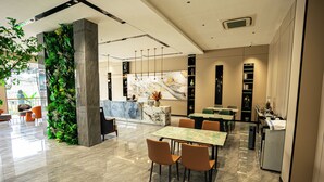 Un hall d’hôtel moderne avec un comptoir d’accueil en marbre, un jardin vertical verdoyant et un espace repas avec des chaises orange et une table en marbre.