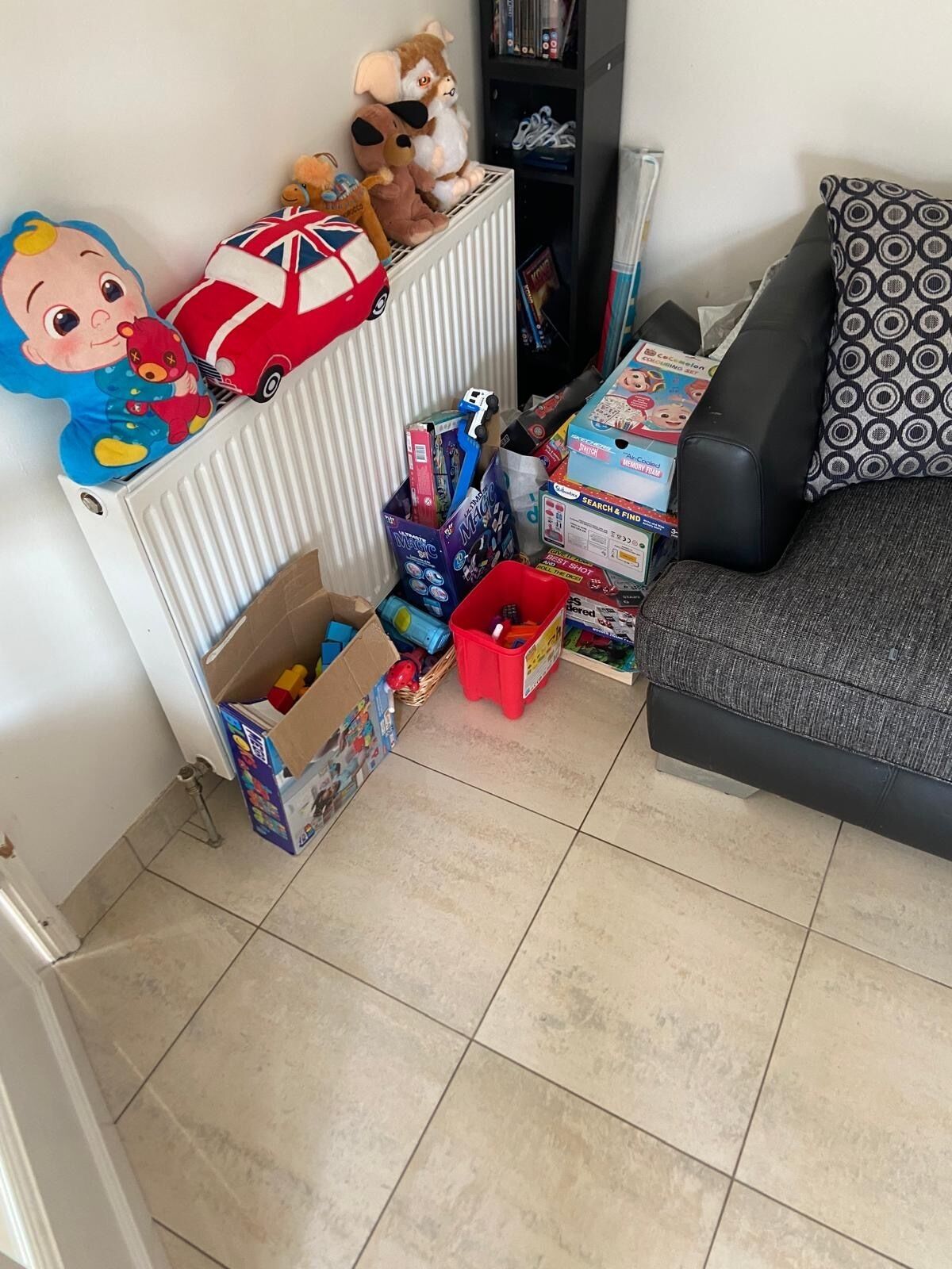 Een rommelige kamer met een radiator, een bank en verschillende speelgoed en dozen.