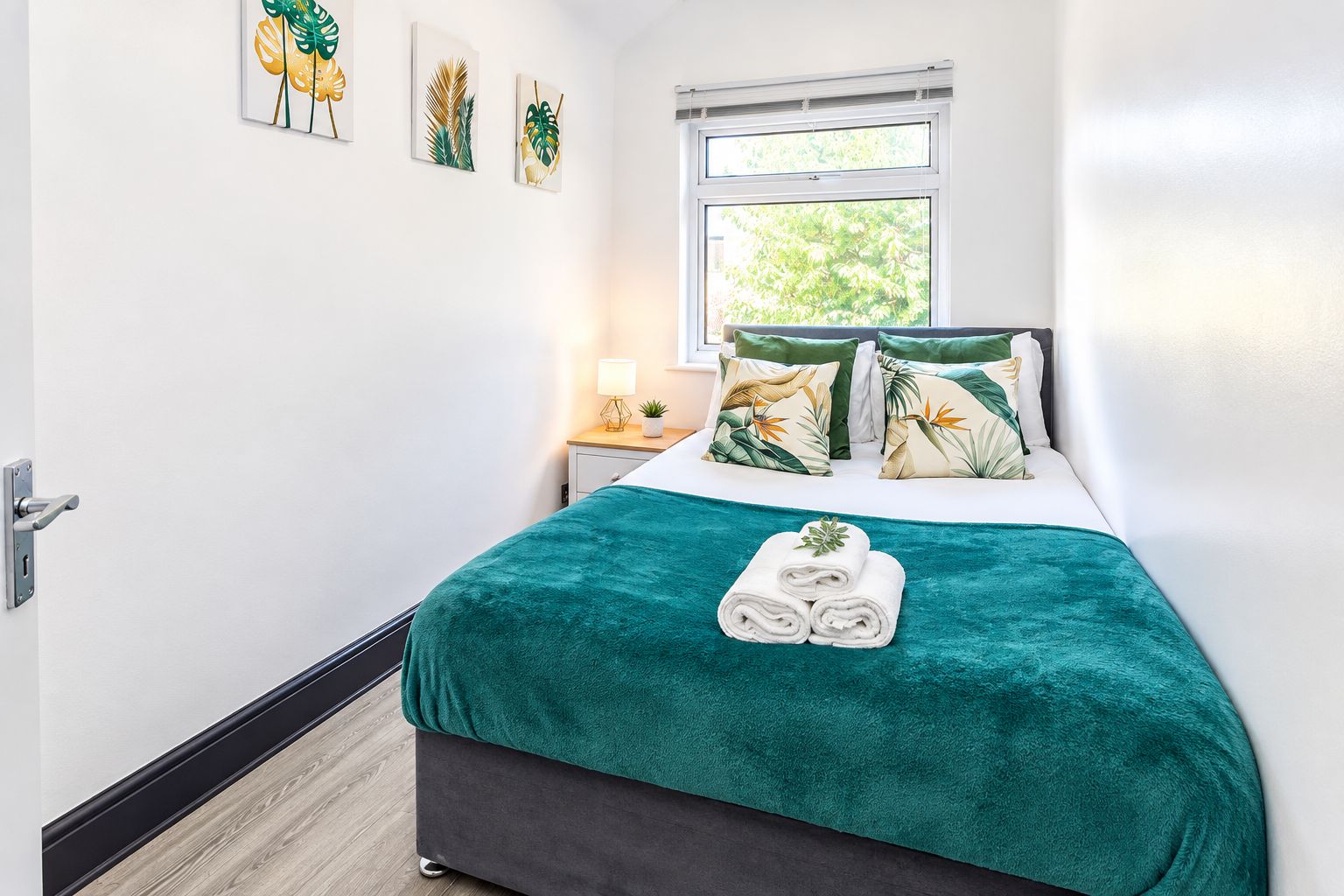 A hotel room with a bed, a window, a nightstand, and wall art.