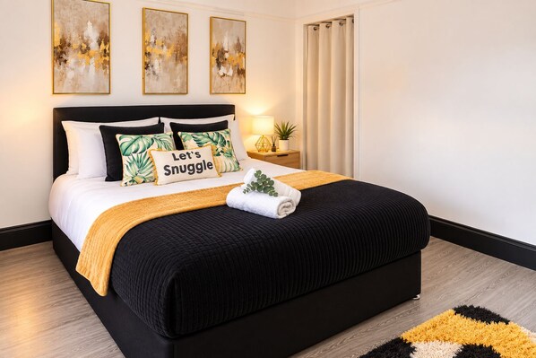 Un dormitorio moderno con una cama negra, almohadas blancas y verdes estampadas, una almohada con la inscripción 'Let's Snuggle', una planta pequeña y una manta amarilla.