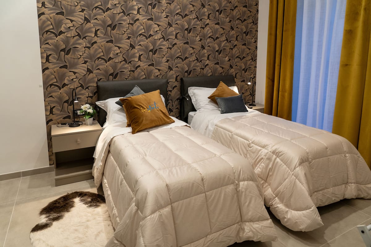 A hotel room with two beds, a patterned wallpaper, a nightstand with a lamp, and a view of a blue sky through curtains.