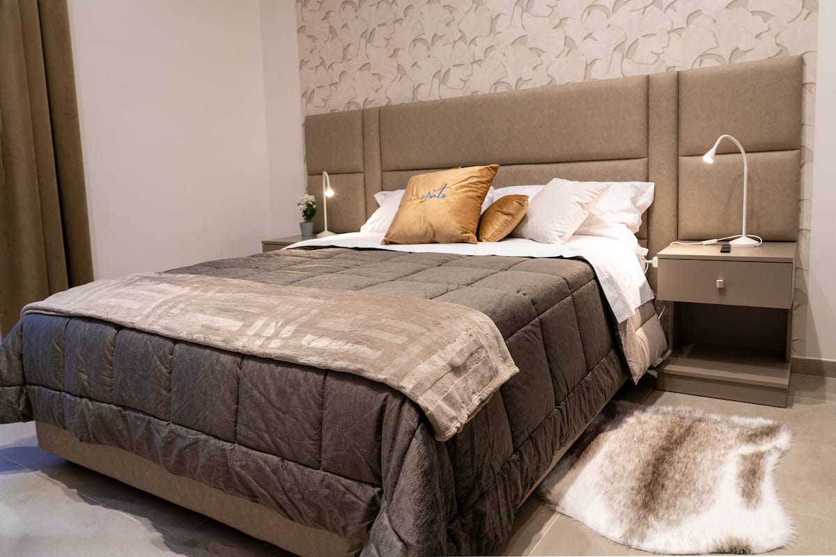 A neatly made bed with a brown quilt, a fluffy rug, and a nightstand with a lamp.