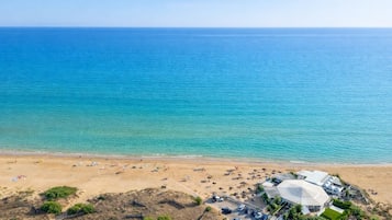 Ein Luftbild zeigt einen Strand mit einem großen, kuppelartigen Gebäude, einem Parkplatz und einem sandigen Strandabschnitt mit Sonnenschirmen und Personen.