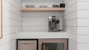 Eine kleine Küche mit weißen Schränken, einem Edelstahlkühlschrank, einer Kaffeemaschine und Dekorationen auf Regalen.