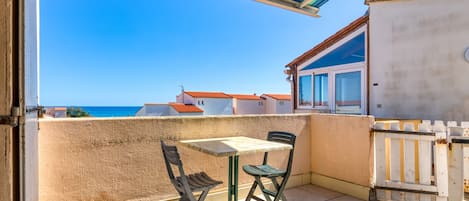 A balcony with a table and chairs, a view of the sea, and a building with a red roof in the background.