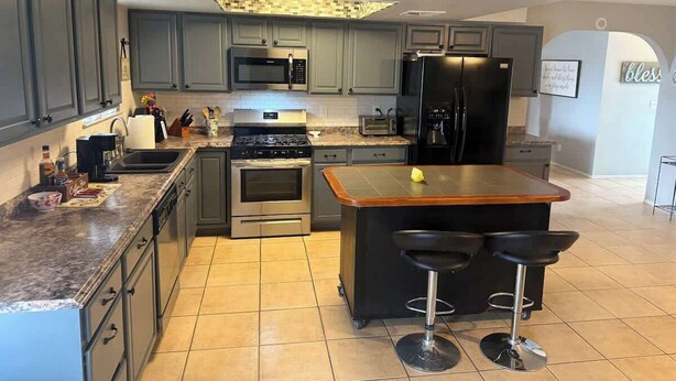 Una cocina moderna con armarios grises, electrodomésticos de acero inoxidable, una isla central con taburetes y piso de baldosas.