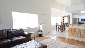 A living room with a leather sofa and ottoman, a dining area with wooden chairs, a kitchen with white cabinets, and a large window providing natural light. - 19th Hole at Hawk Ridge (Mesquite)