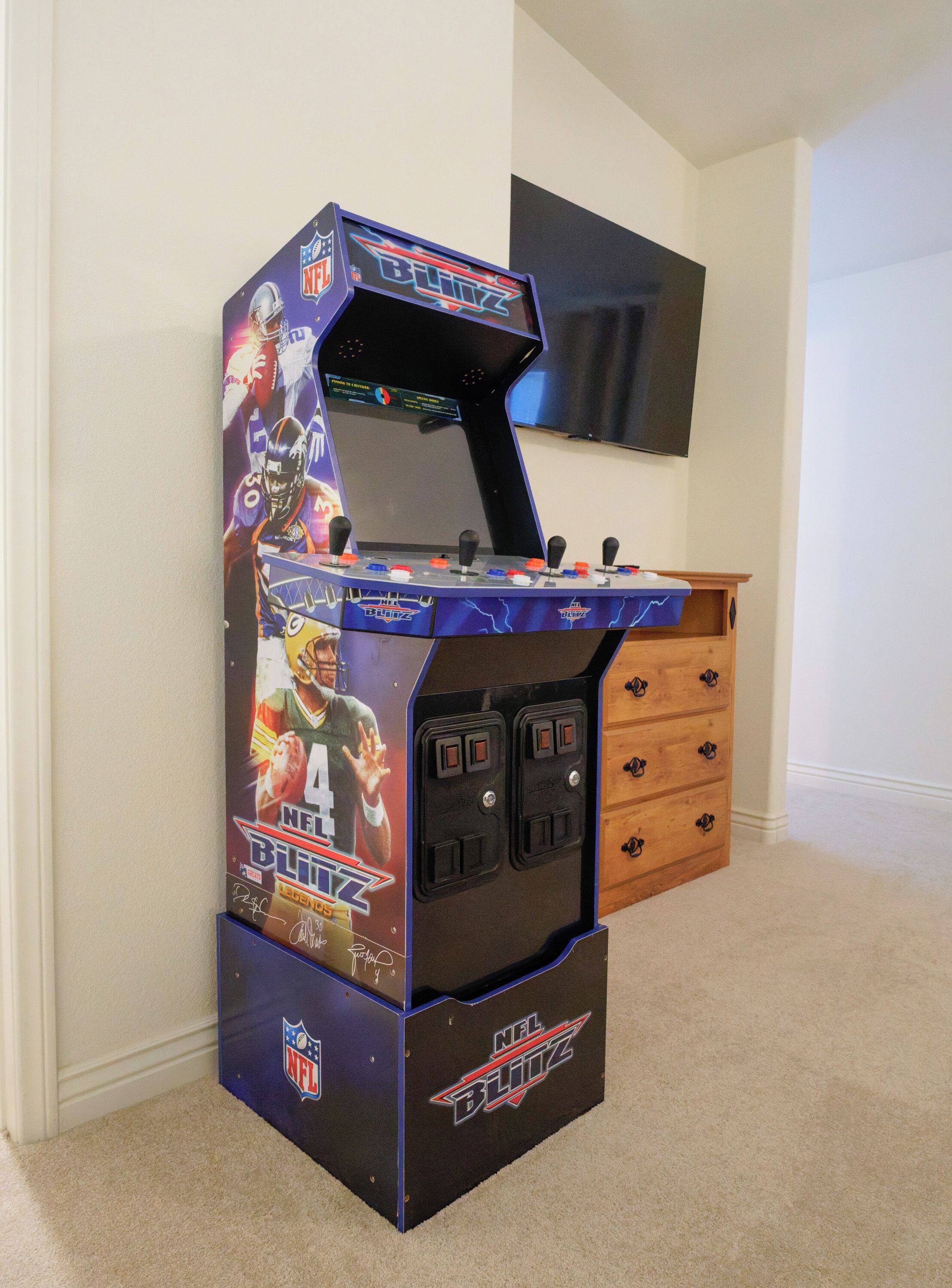 A gaming arcade machine with NFL Blitz branding, featuring a large screen and multiple joysticks, placed in a room with a wooden cabinet and a television on the wall.