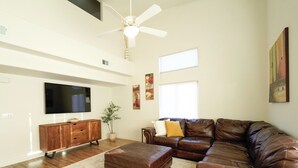 A spacious living room with a large leather sofa, a wooden entertainment unit, a flat-screen TV, a ceiling fan, and a potted plant. - 19th Hole at Hawk Ridge (Mesquite)