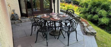 An outdoor patio with a round table and chairs, a water fountain, and a view of a garden and trees.