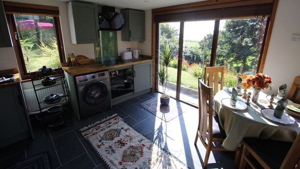 A kitchen with a washing machine, a dining area with a table set for two, and a view of the garden through the sliding glass door. - The Rookery Sandside-Stylish Luxury Home (Kirkby in Furness)