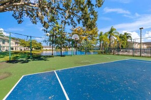 Quadra de basquete ao ar livre com superfície azul, cercada por uma cerca verde, árvores e um corpo de água ao fundo.