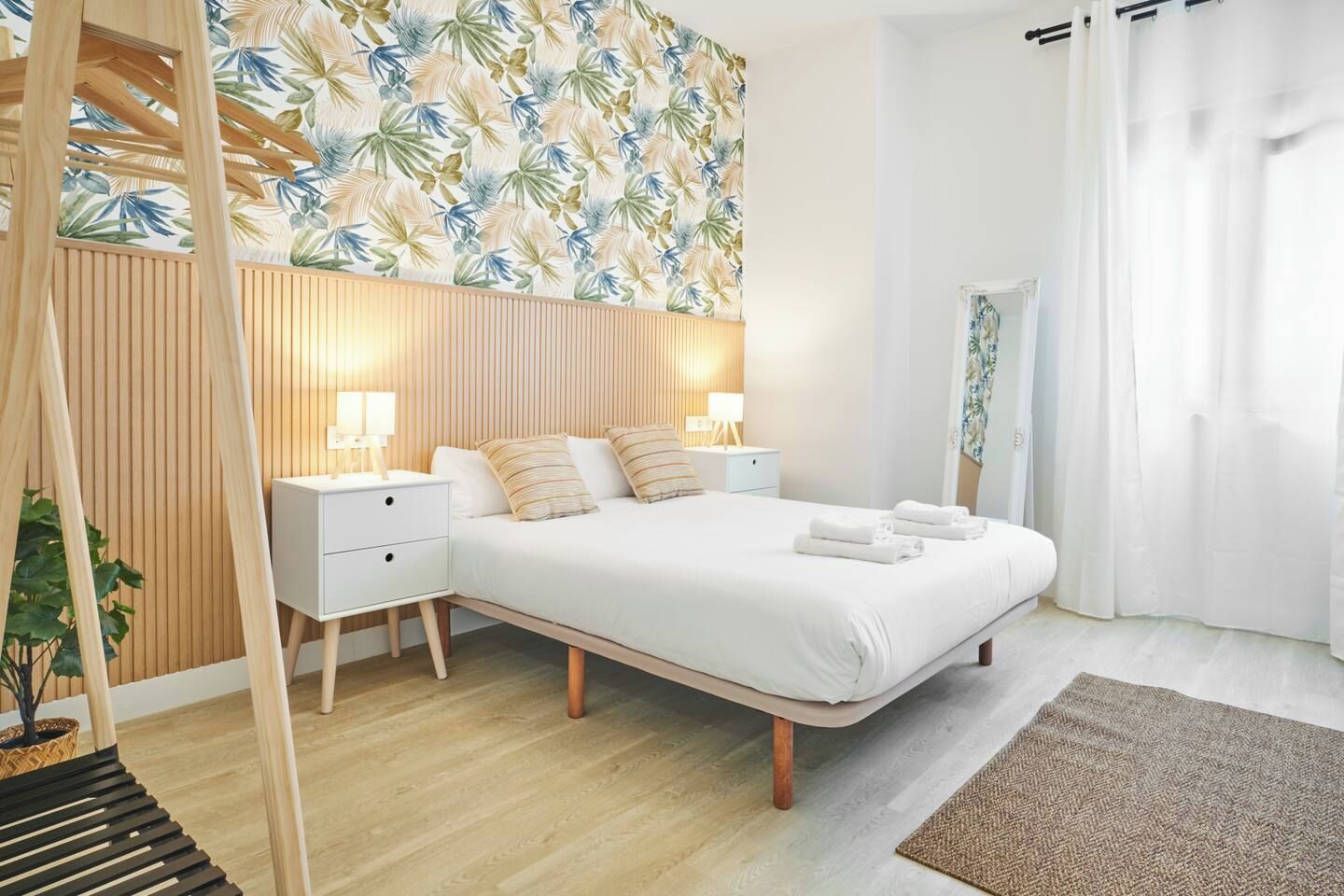 Un dormitorio moderno con una cama grande, mesitas de noche, un cabecero de madera y un papel tapiz con estampado de hojas tropicales.