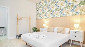 A modern bedroom with a large bed, two bedside tables, a wooden headboard, and a tropical leaf pattern on the wall.