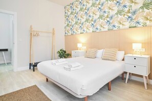 Un dormitori modern amb un llit gran, dues taules de nit, un panell de fusta al capçal i un motiu de fulles tropicals a la paret.