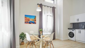 A modern kitchen with a washing machine, a dining area with a glass table and chairs, and a window with curtains.