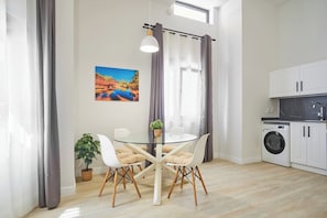 Cuina moderna amb rentadora, zona de menjador amb taula de vidre i cadires, i finestra amb cortines.