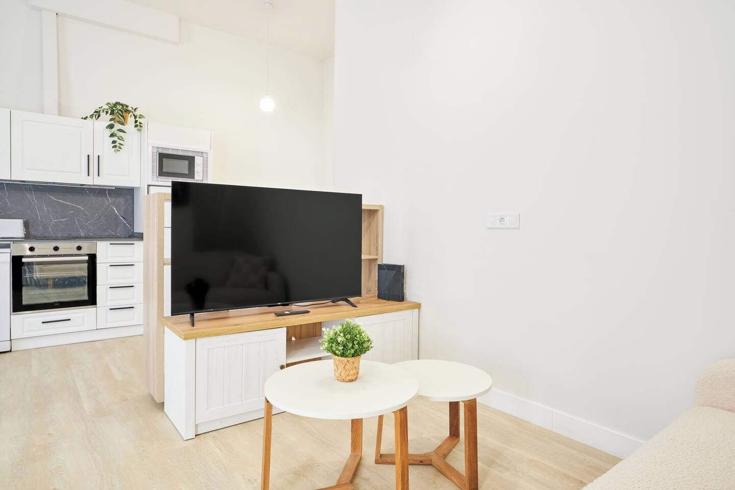 Cocina moderna con horno integrado, microondas y un televisor de pantalla plana. Hay dos mesas blancas con patas de madera y una pequeña planta en maceta sobre la encimera.