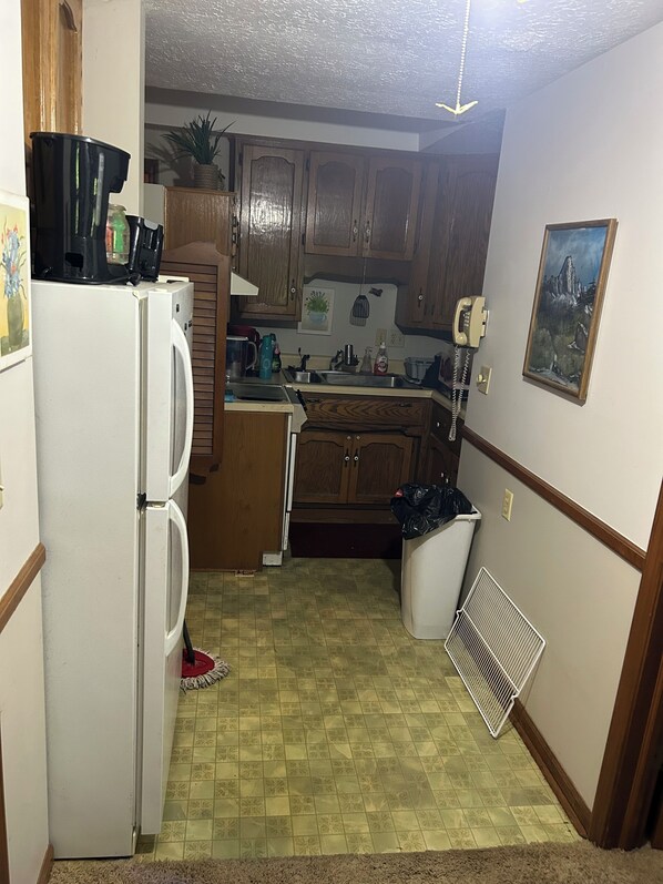 A compact kitchen with wooden cabinets, a refrigerator, and a microwave. There is a trash bin and a vent near the sink.