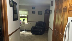 Un pasillo con piso alfombrado, ventilador de techo, sillón de cuero negro y refrigerador blanco.