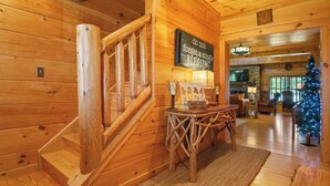 A wooden cabin interior with a staircase, a 'Welcome Cabin' sign, a Christmas tree, and a fireplace.