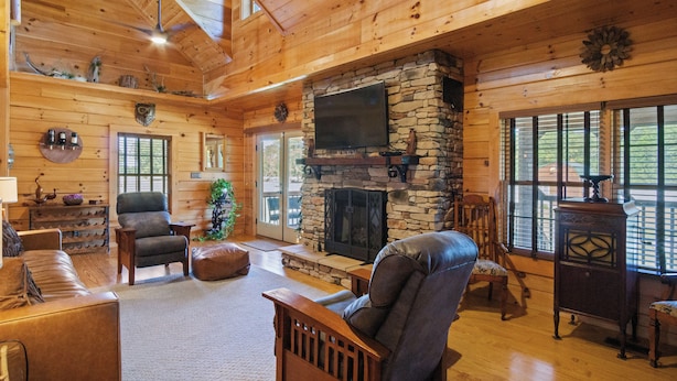 A spacious living room with a stone fireplace, wooden ceiling, and large windows.