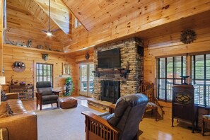 A spacious living room with a stone fireplace, wooden ceiling, and large windows.