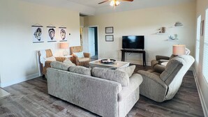 Uma sala de estar com sofá, poltronas, mesa de centro, televisão e ventoinha no teto.
