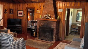 A cozy living room with wooden walls, a fireplace, a television, and a bookshelf. - Charming Cabin 500 feet to beach (Plymouth)