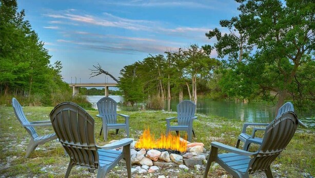 A fire pit with four Adirondack chairs around it, set on a gravel area near a body of water with trees and a bridge in the background. - Guadalupe Riverfront Retreat | Sleeps 10 Kerrville (Kerrville)