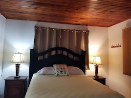 Quarto com teto de madeira, uma cama com cabeceira, duas mesas de cabeceira com luminárias e uma cortina.