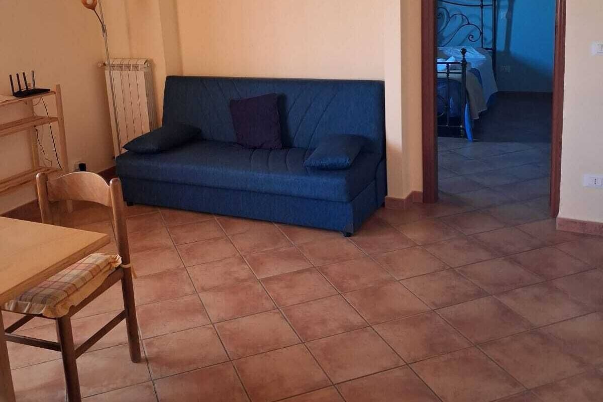 A room with a blue sofa, a wooden dining table, and a chair. There is a radiator on the wall and a door leading to another room.