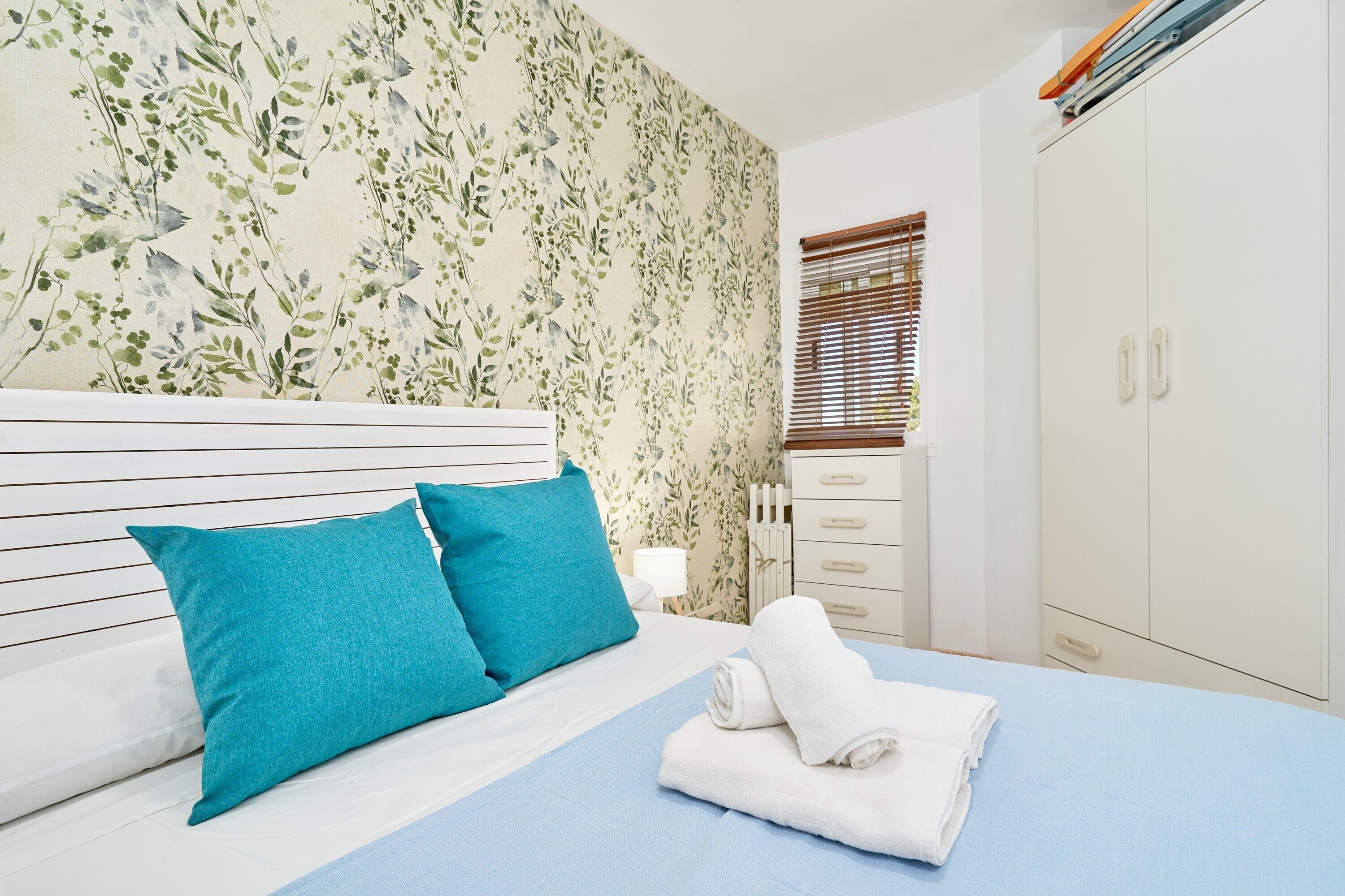 A bedroom with a bed, two pillows, a nightstand, a lamp, a wardrobe, and a window with blinds.