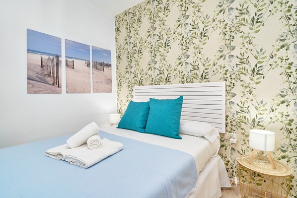 Una cama bien hecha con ropa de cama azul, un papel tapiz floral y un cuadro colgado en la pared de una playa.