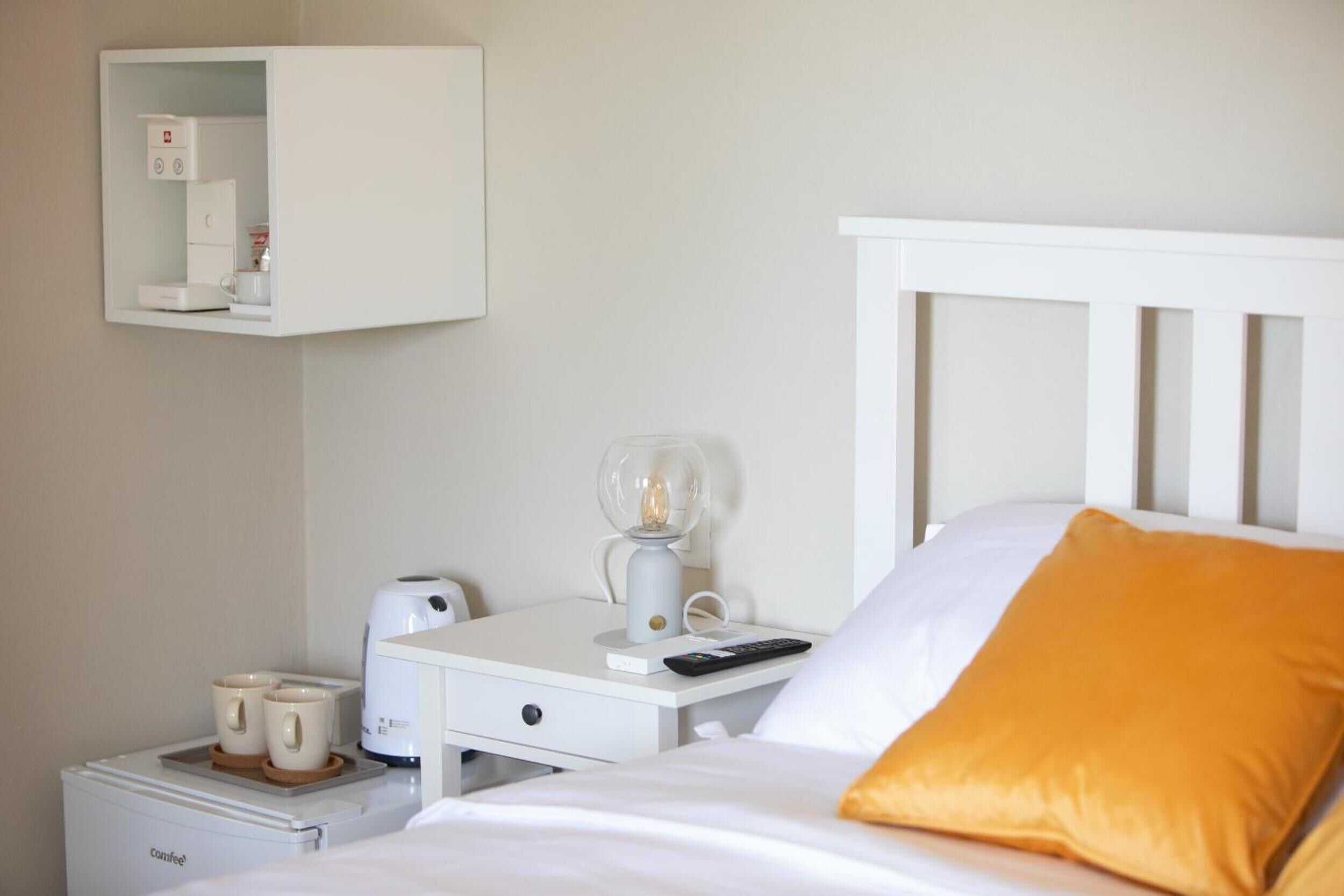 A hotel room with a bed, a bedside table with a coffee maker, a lamp, and a small cabinet.