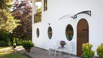 Een wit gebouw met boogvensters, een deur en een balkon met een hekwerk. Er staan potplanten en een stoel buiten.
