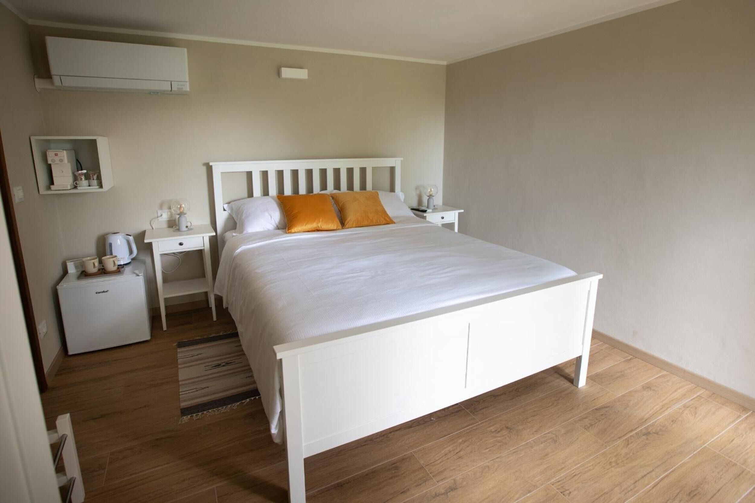 Une chambre avec un lit blanc, deux tables de chevet, une petite armoire blanche et une machine à café.