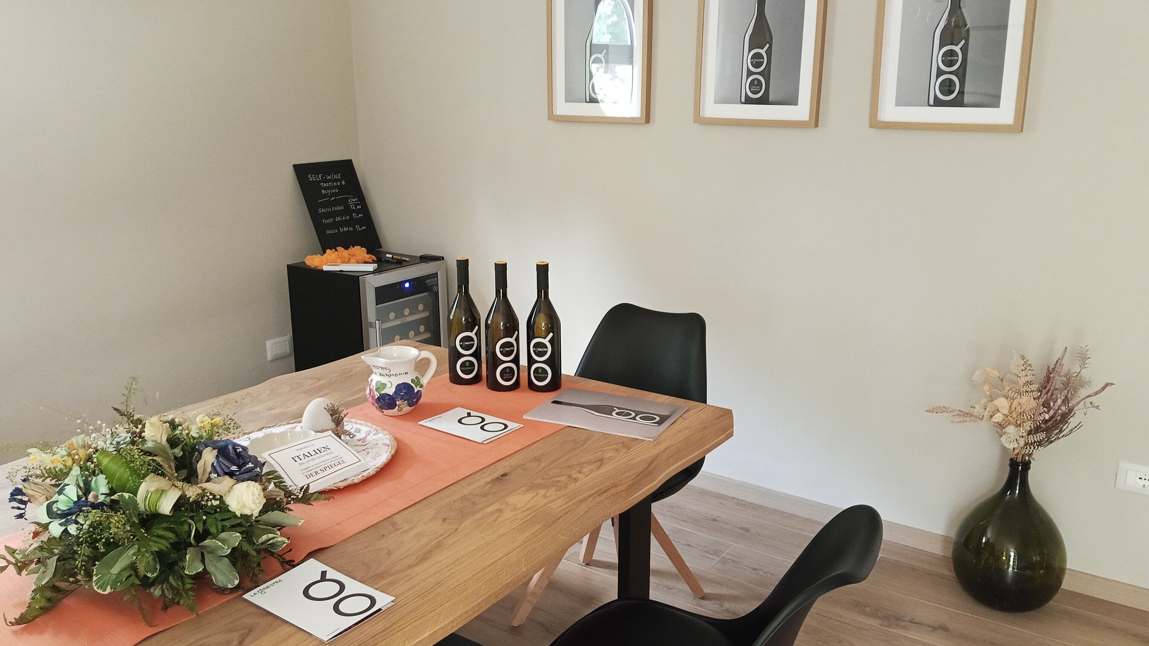 Una mesa de comedor con botellas de vino, una jarra y un menú. Hay cuadros en la pared y un florero con flores secas.