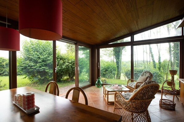 A dining area with a wooden table, wicker chairs, and a view of greenery through large windows.
